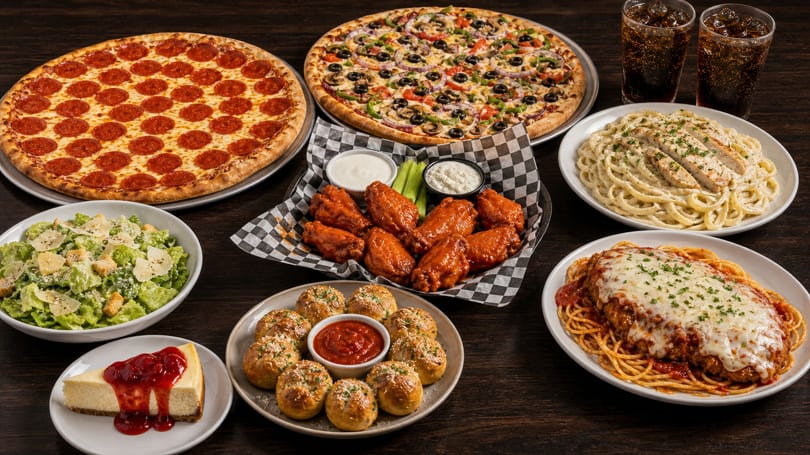 A feast of two pizzas, wings, pasta dishes, salad, garlic knots, cheesecake, and sodas.