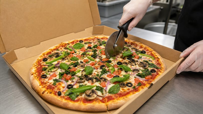 A gloved hand uses a pizza cutter to slice a fresh vegetable pizza in a cardboard box.